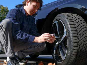 Person crouching beside a black car, checking a tire on an alloy wheel outdoors on a sunny day with trees and fallen leaves nearby.