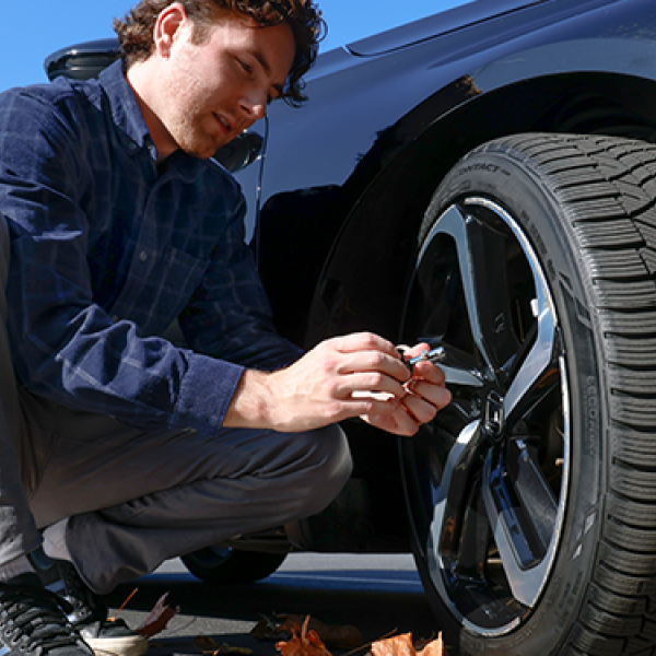 Person crouching beside a black car, checking a tire on an alloy wheel outdoors on a sunny day with trees and fallen leaves nearby.